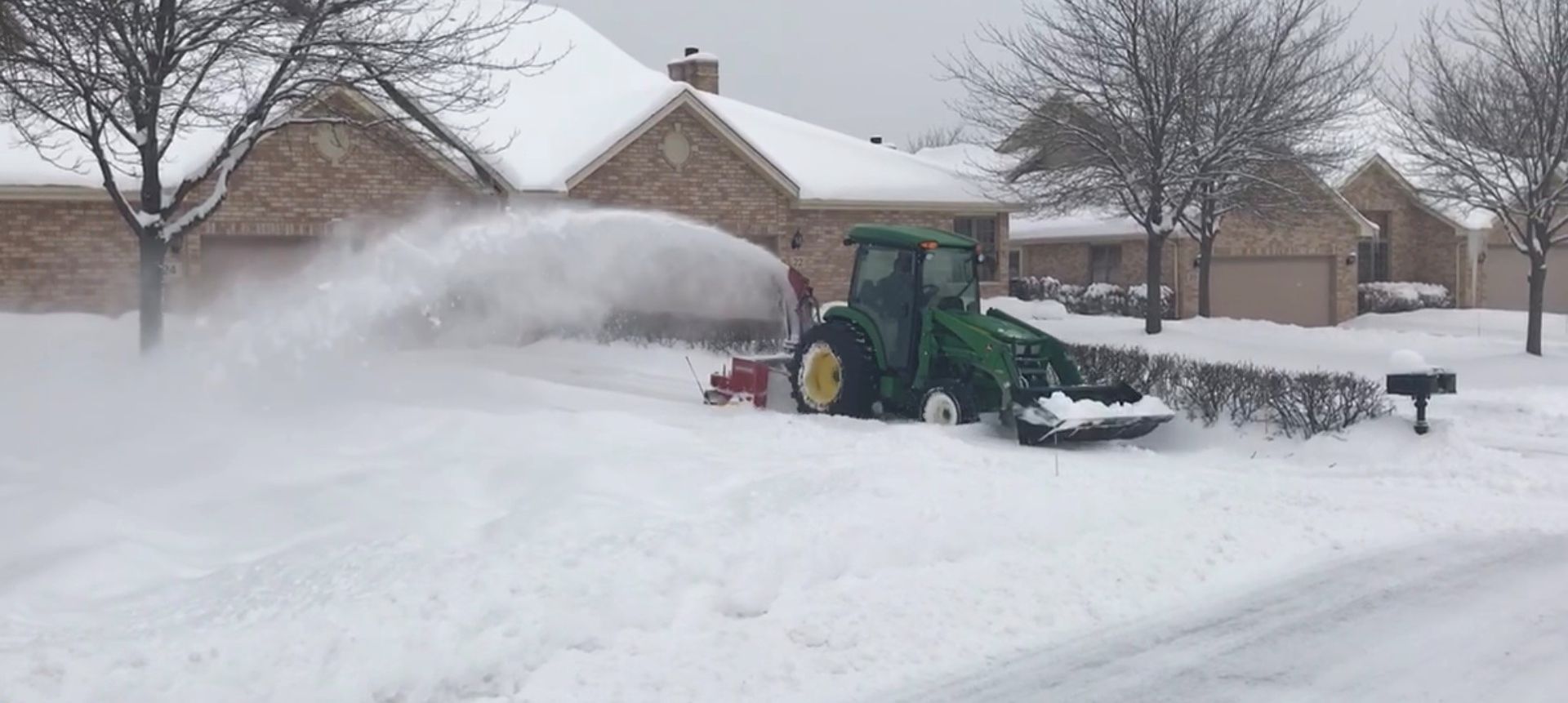 A John Deere tractor blowing snow in front of houses in a snowy neighborhood.