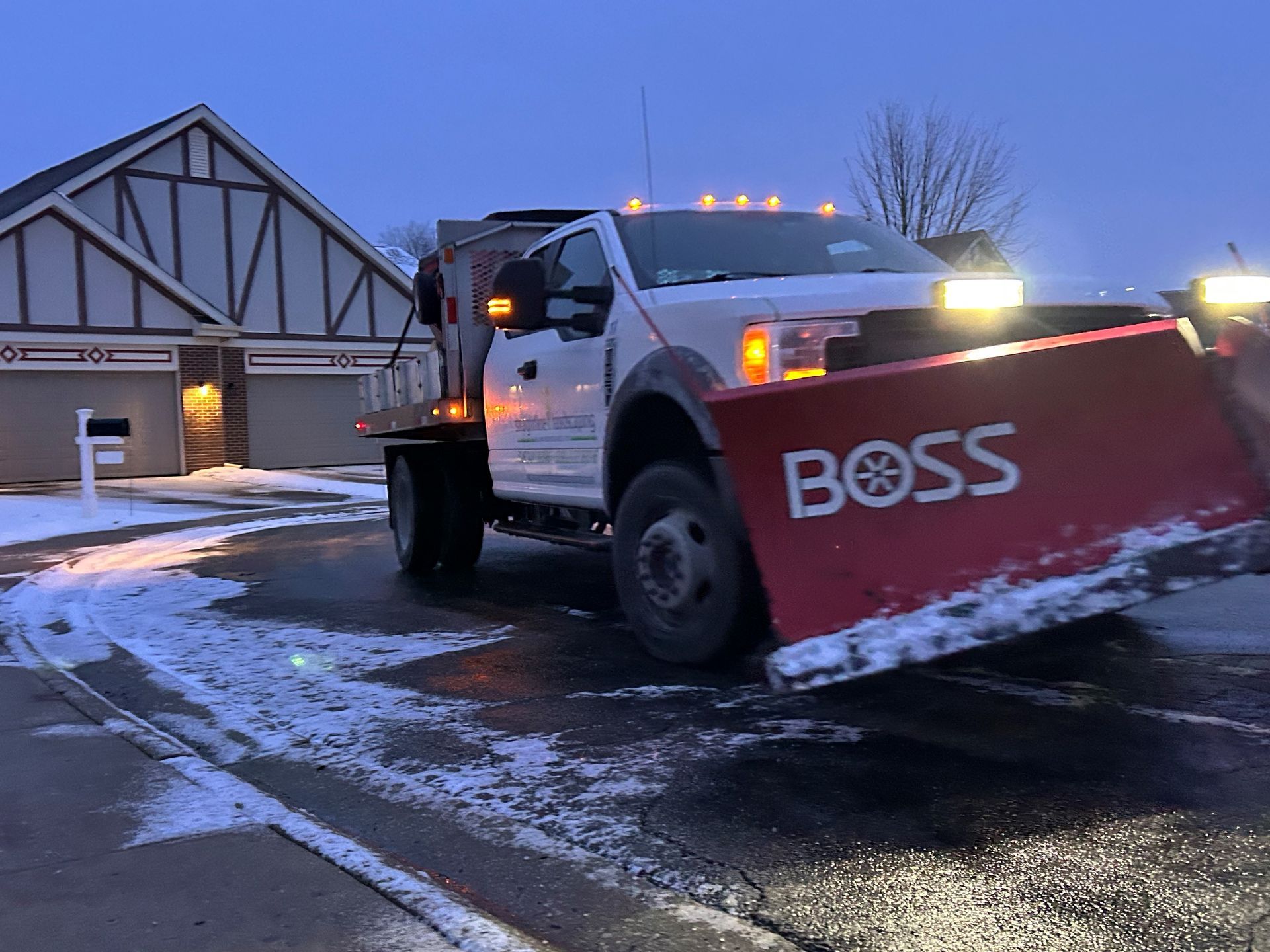 Snowplow truck plowing a residential driveway at dusk. Red plow blade, white truck, snow-covered road.