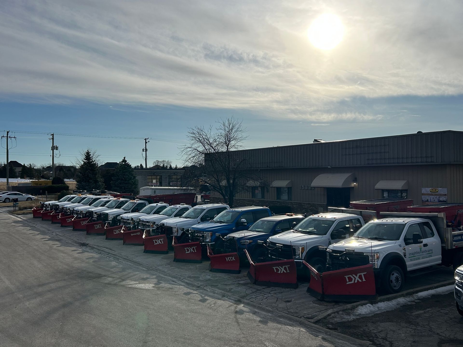 Line of snowplow trucks parked on a snowy lot under a sunny sky.