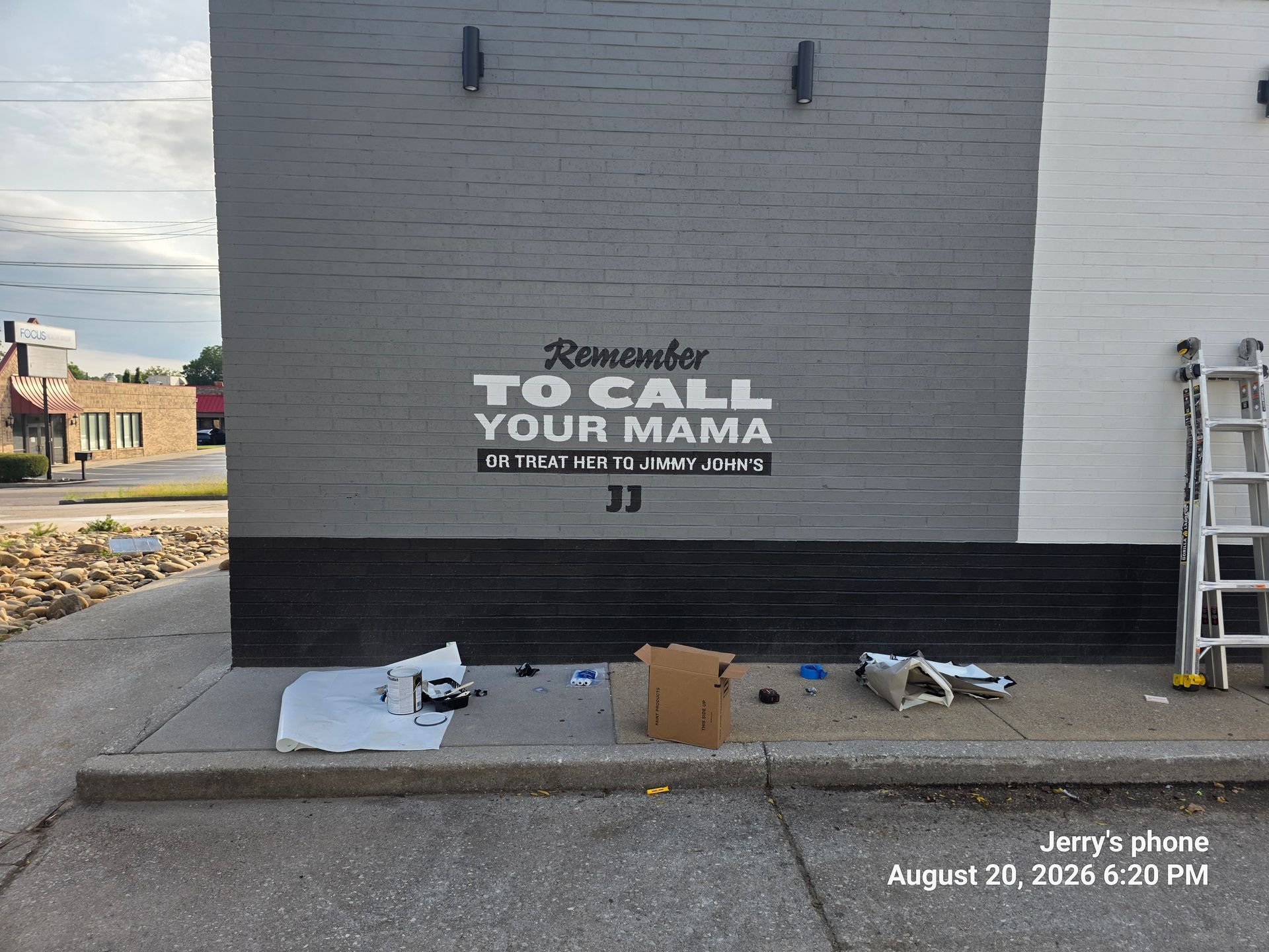 Person spray-painting “TO CALL YOUR MAMA” on a gray brick wall; supplies on sidewalk near ladder