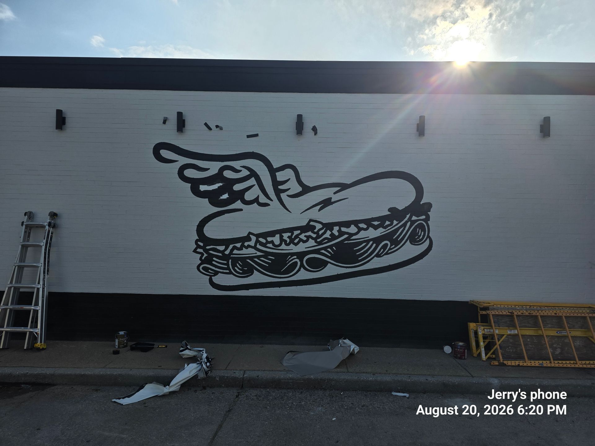 Black-and-white mural of a hand pointing over a sandwich on a wall, with a ladder and tools below