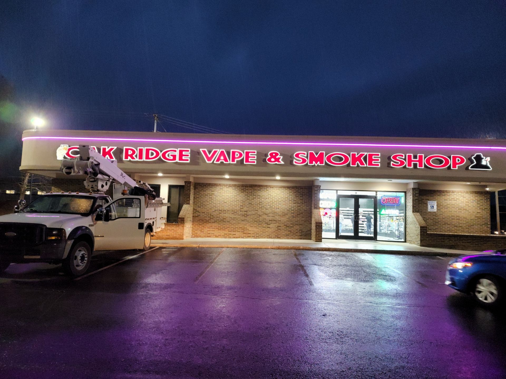 Night exterior of a vape and smoke shop with lit pink signage, wet parking lot, and parked vehicles