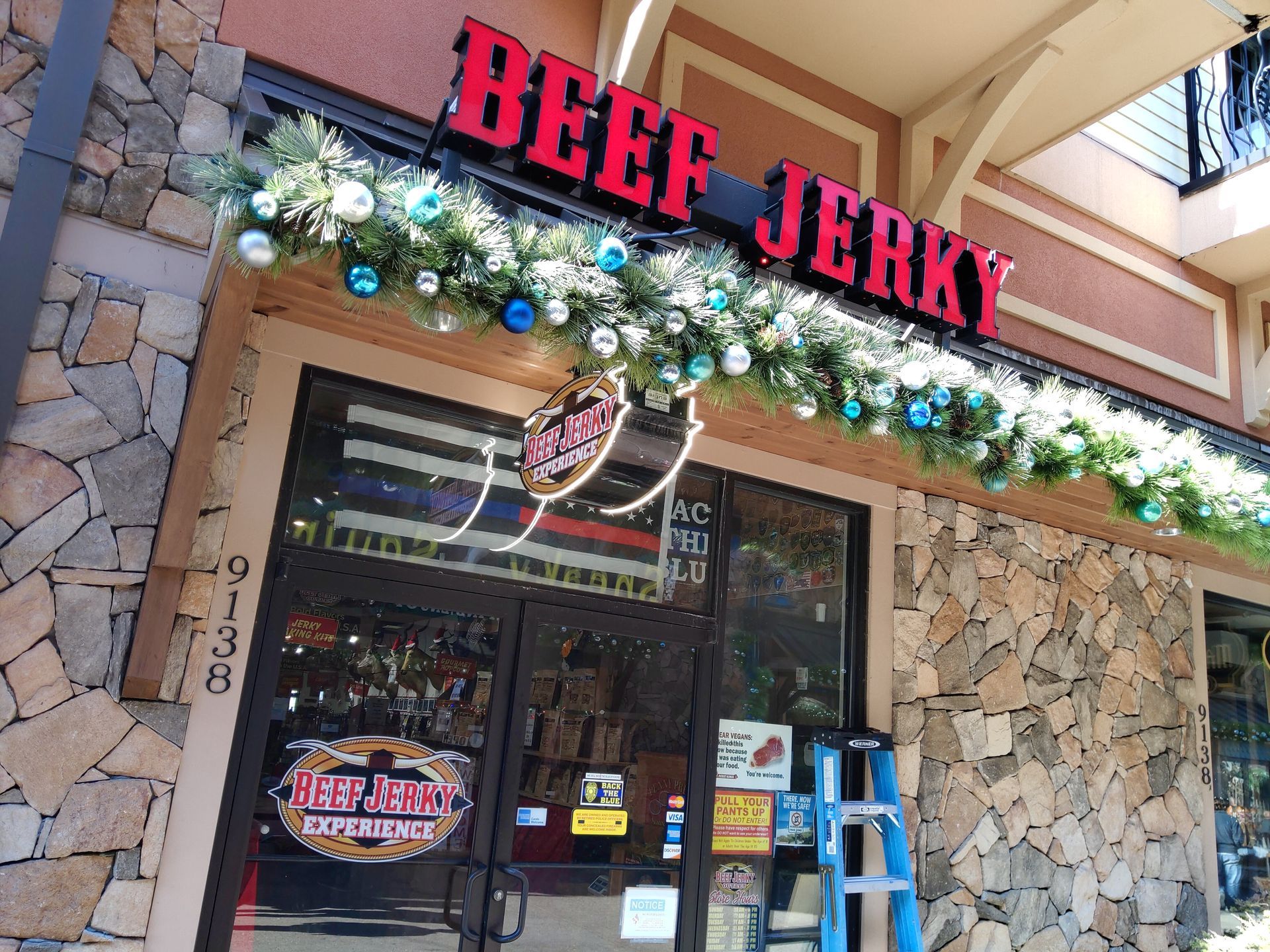 Beef Jerky storefront with red sign, stone facade, holiday garland, and blue ladder by the entrance