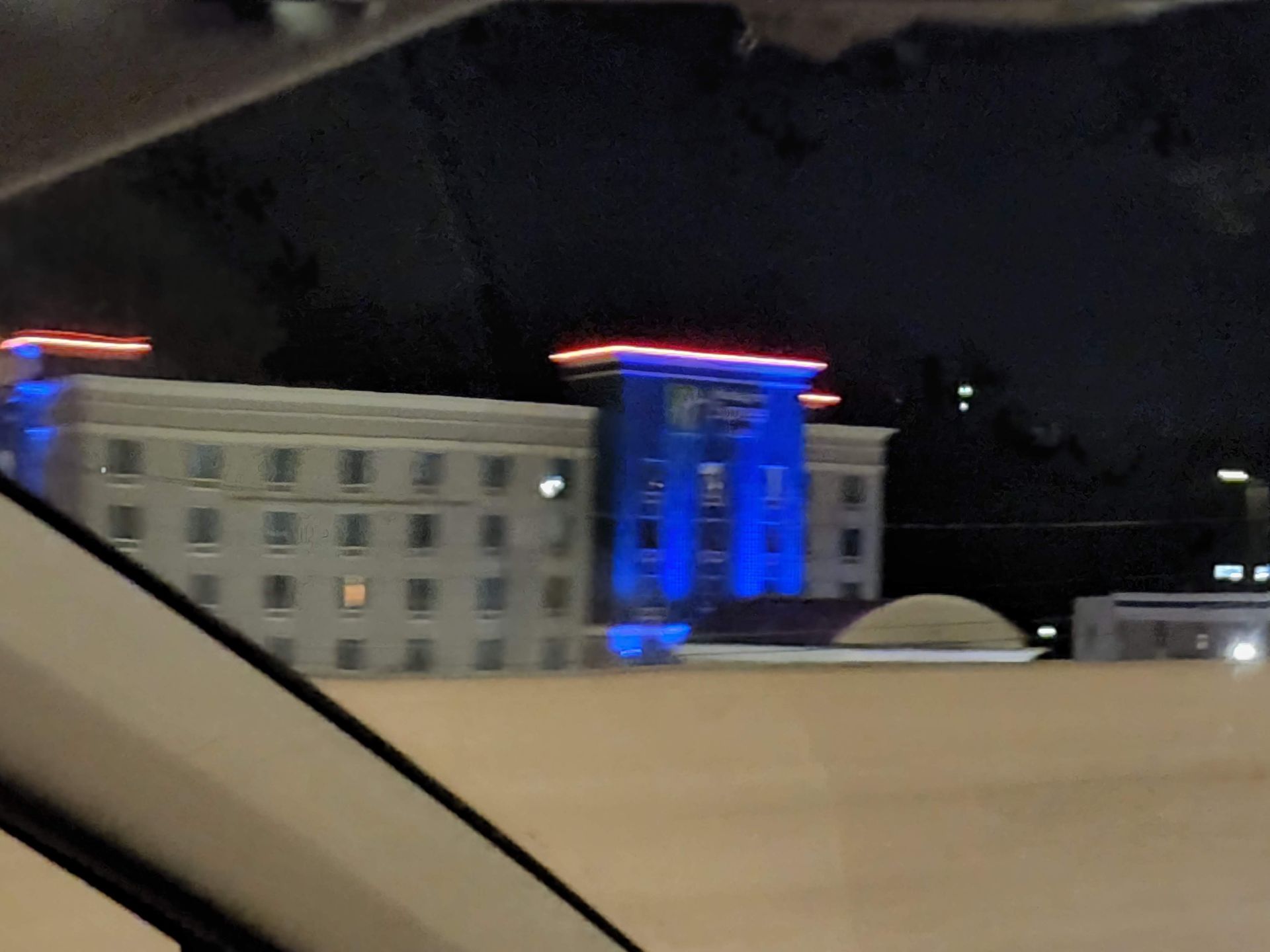 Nighttime city view from a car, with a blue-lit building and a beige barrier in the foreground