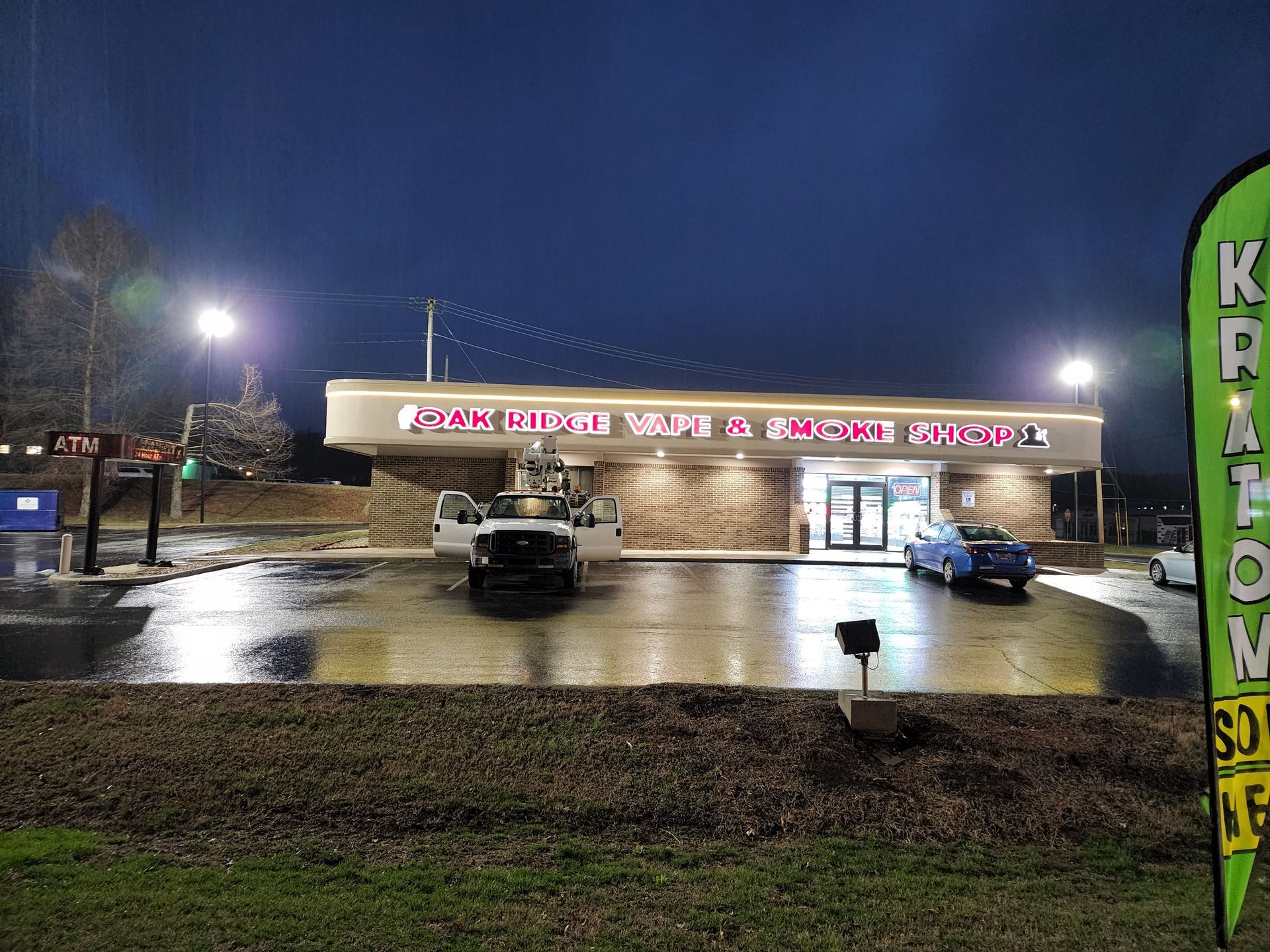 Nighttime storefront with wet parking lot, parked cars, and a bright pink sign.