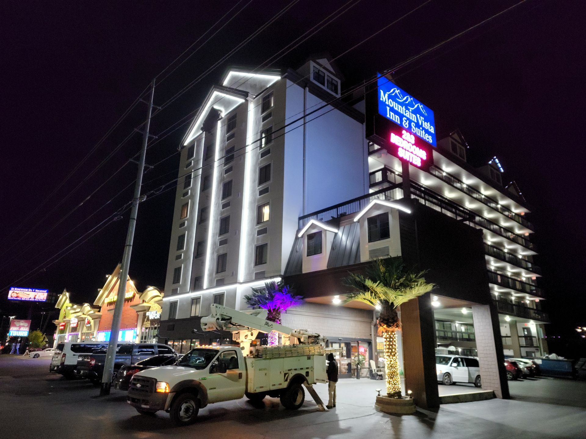 Nighttime street scene with a lit hotel building, parked vehicles, and a busy sidewalk.