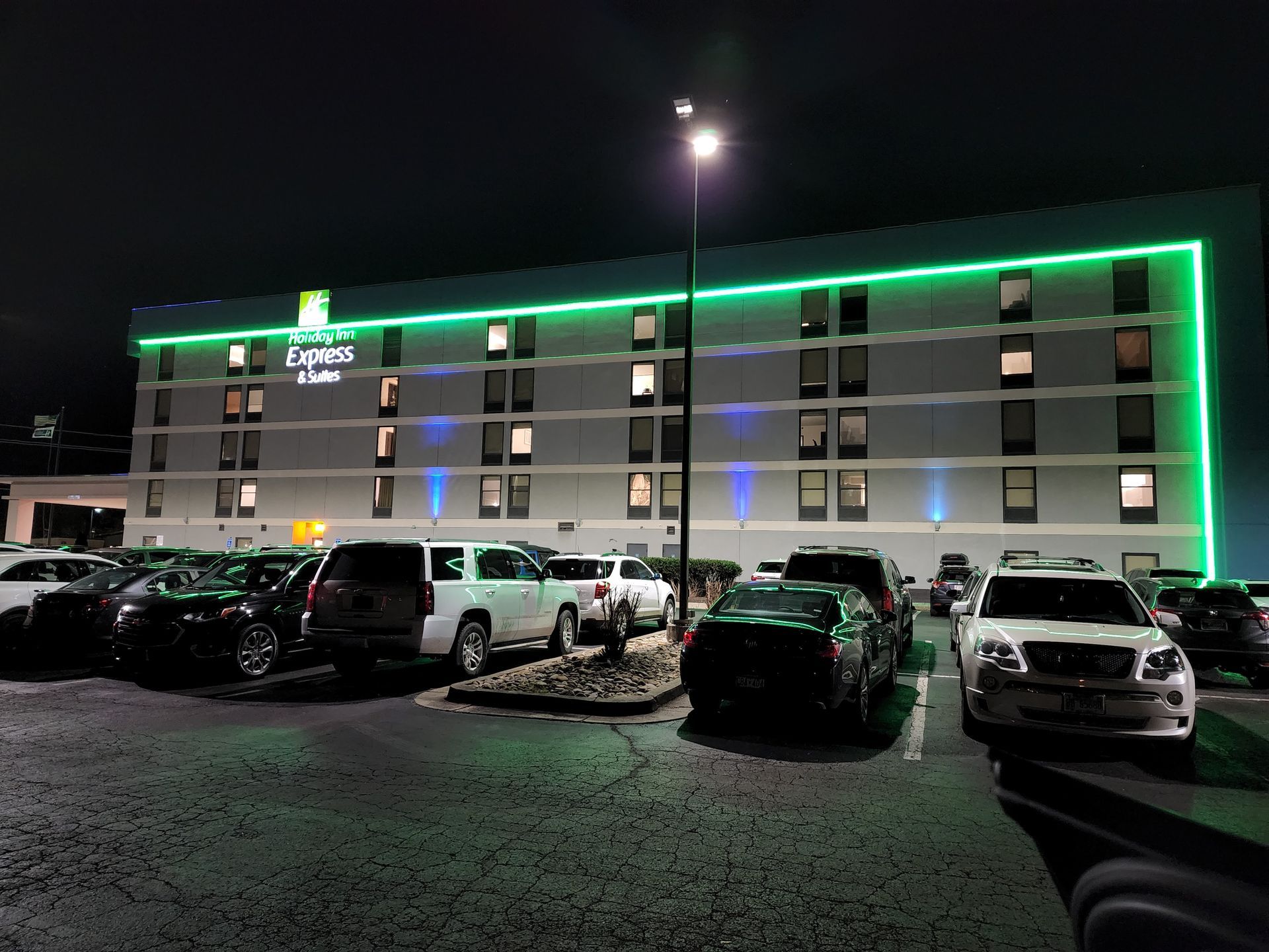 Night view of a lit hotel with green roof lights and parked cars in front.
