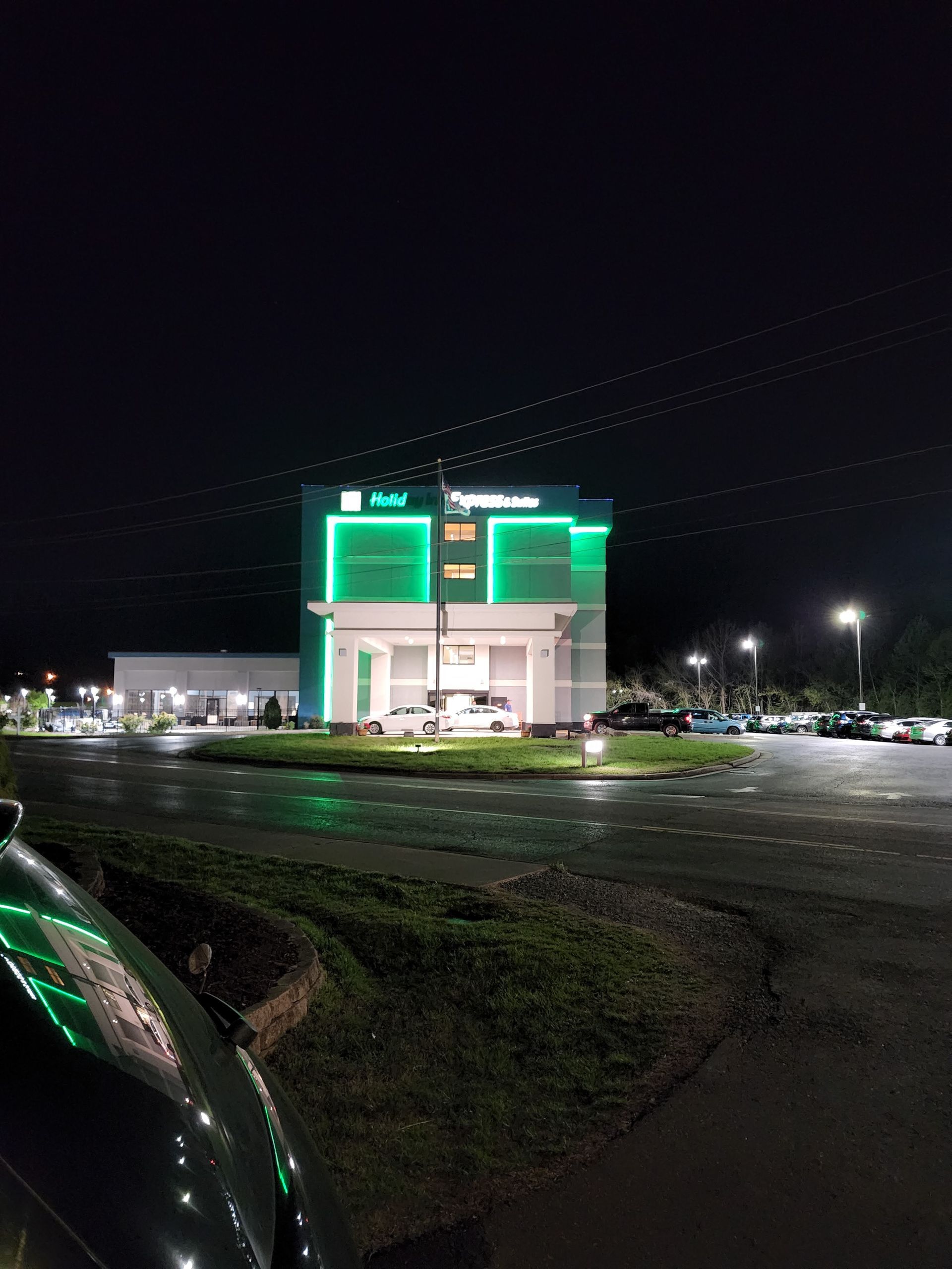 Nighttime view of a lit green storefront and parking lot beside a road
