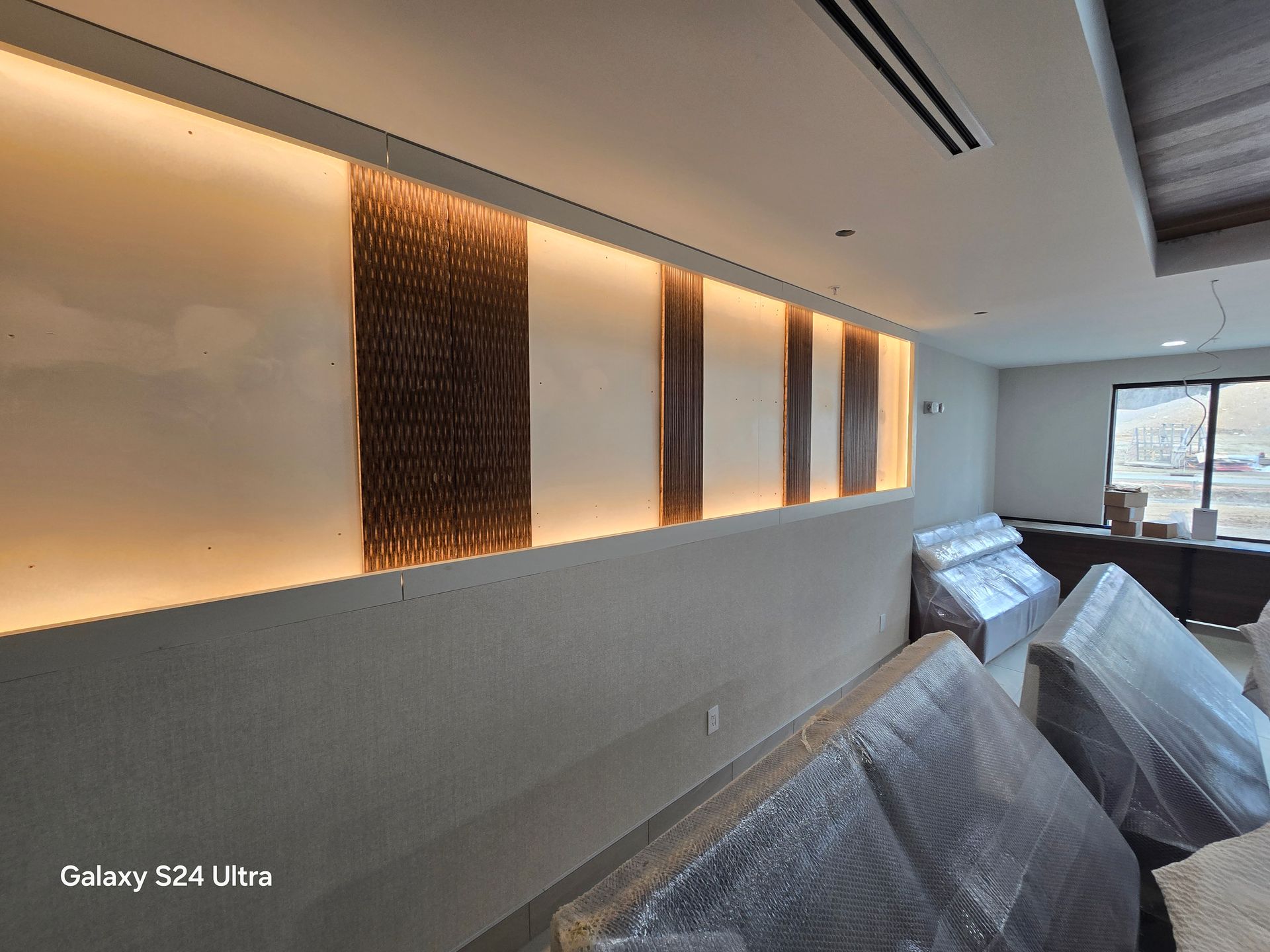 Modern room with wall lighting, textured panels, and furniture covered in plastic sheeting near a window.