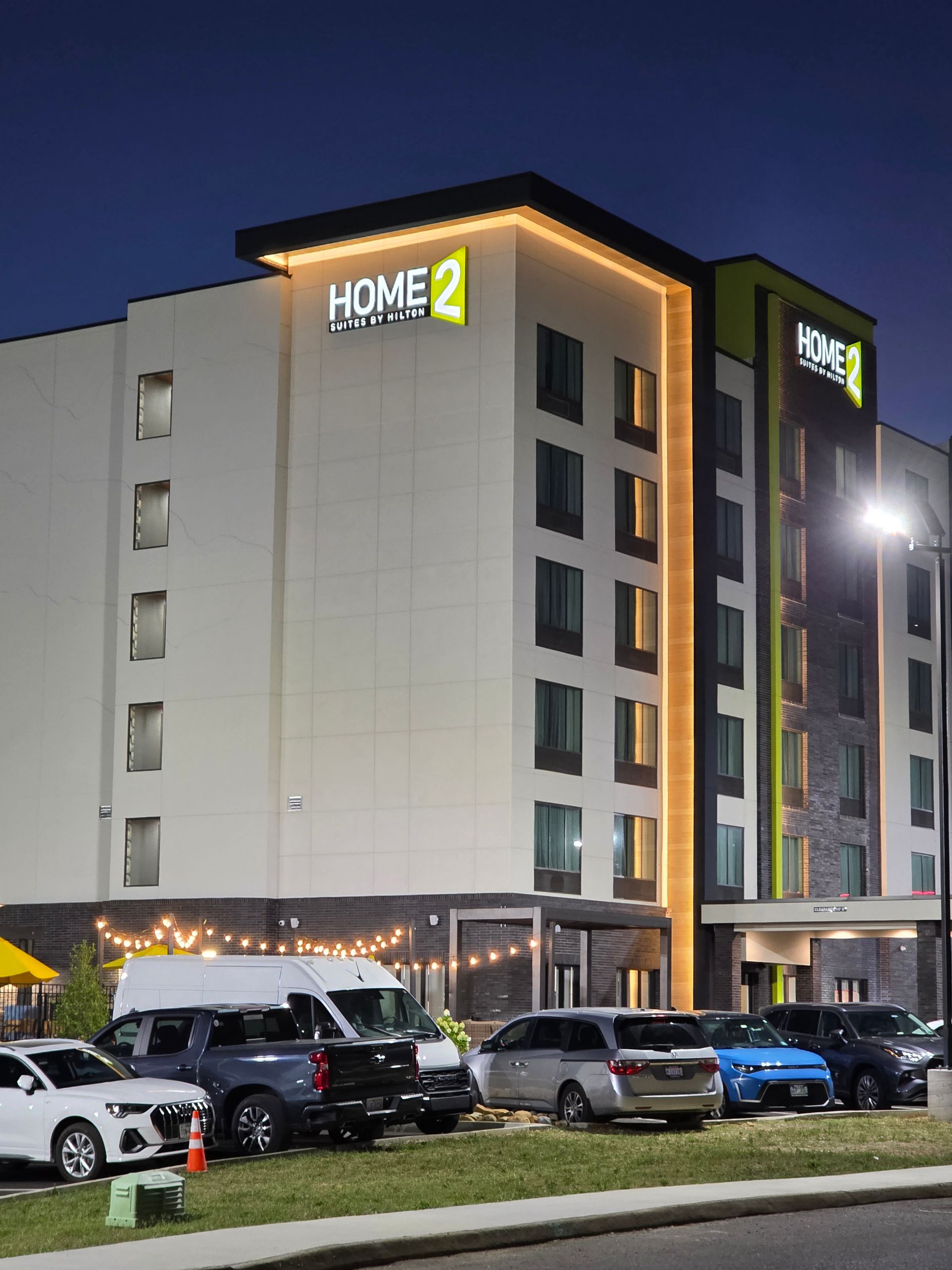 Home2 Suites hotel at dusk with parked cars in front and illuminated signage.