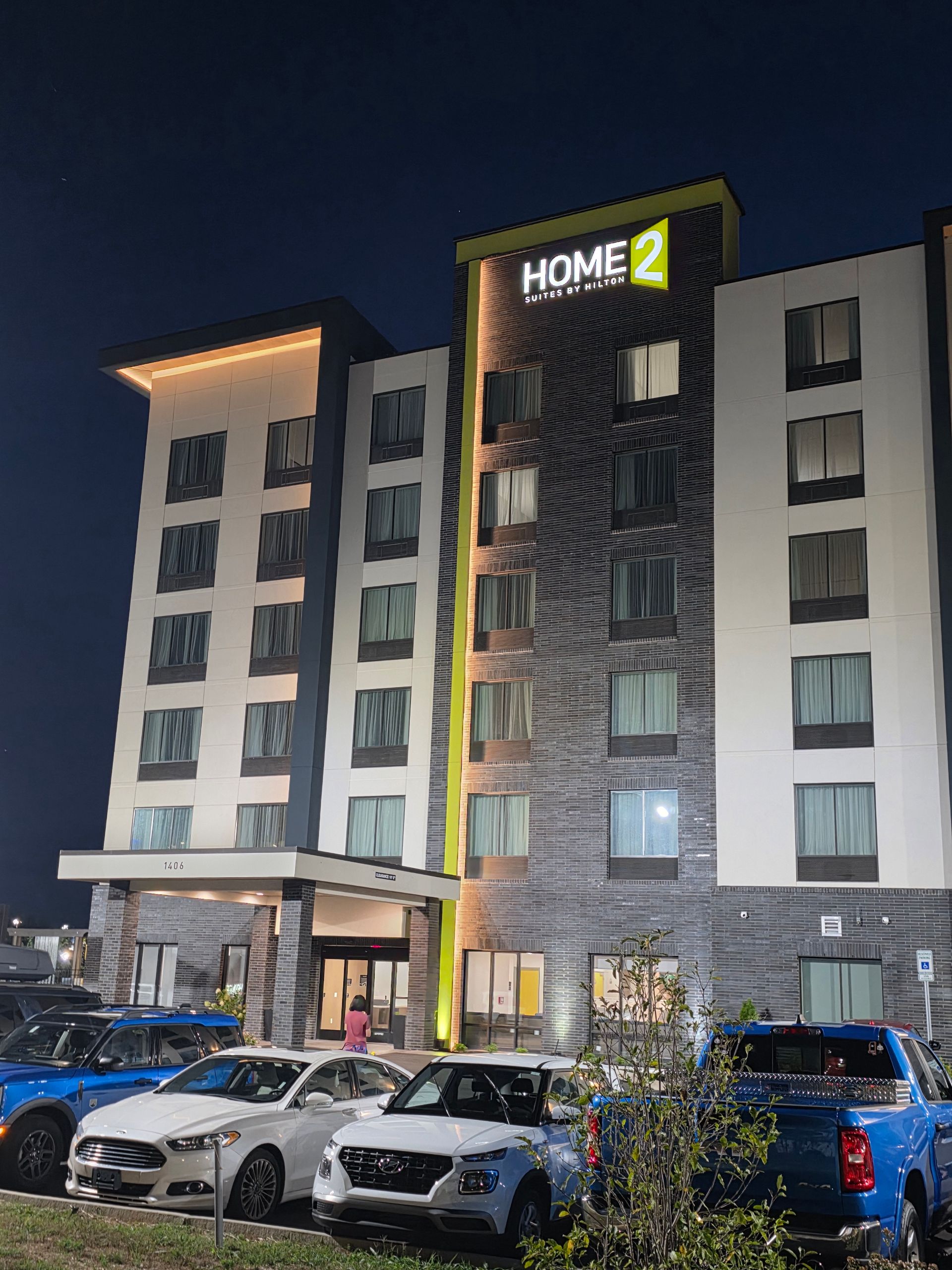 Nighttime exterior of a modern hotel with parked cars in front and illuminated signage on the facade
