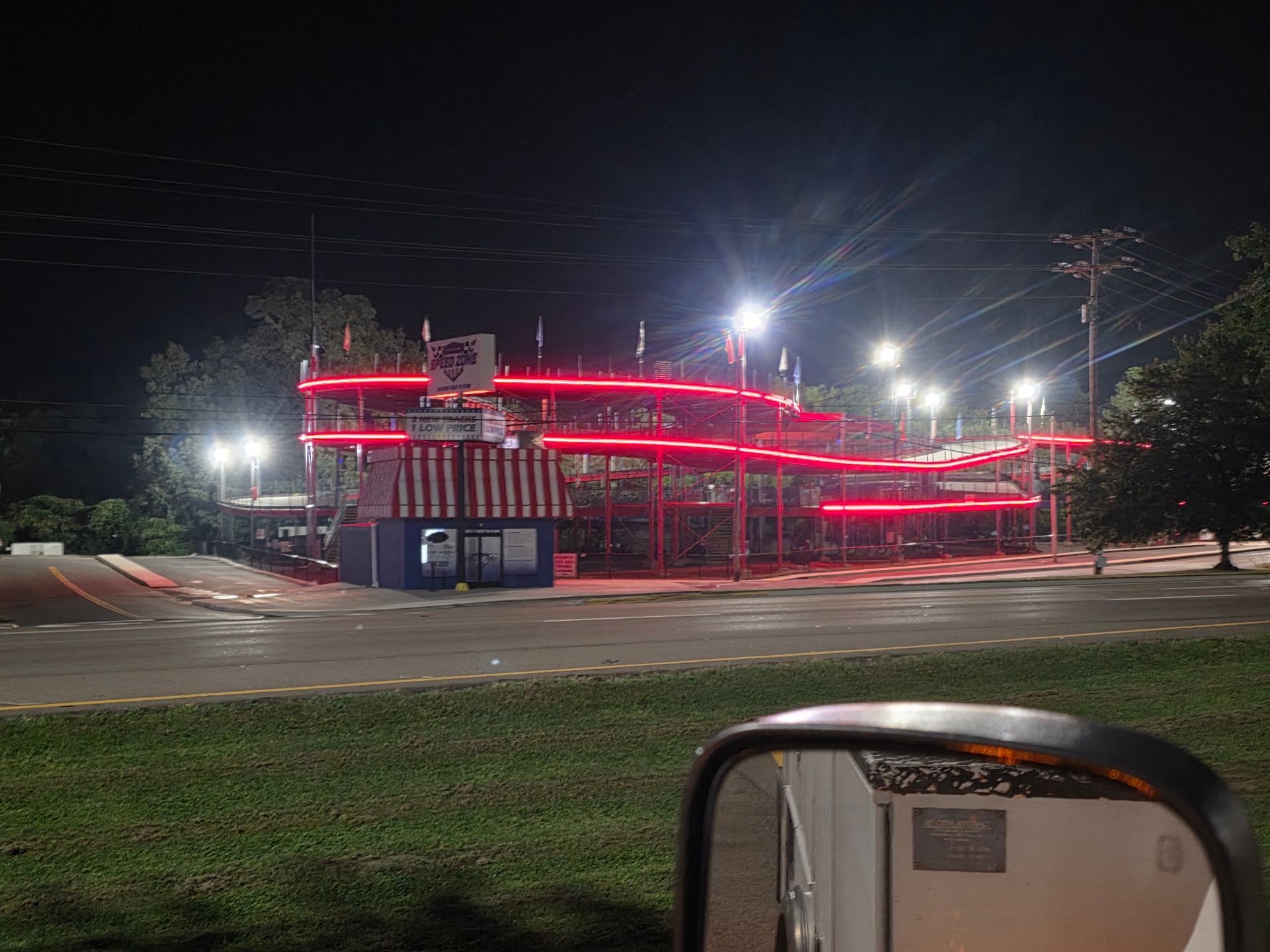 Night view of a red neon-lit building across a road, with grass and a car mirror in the foreground