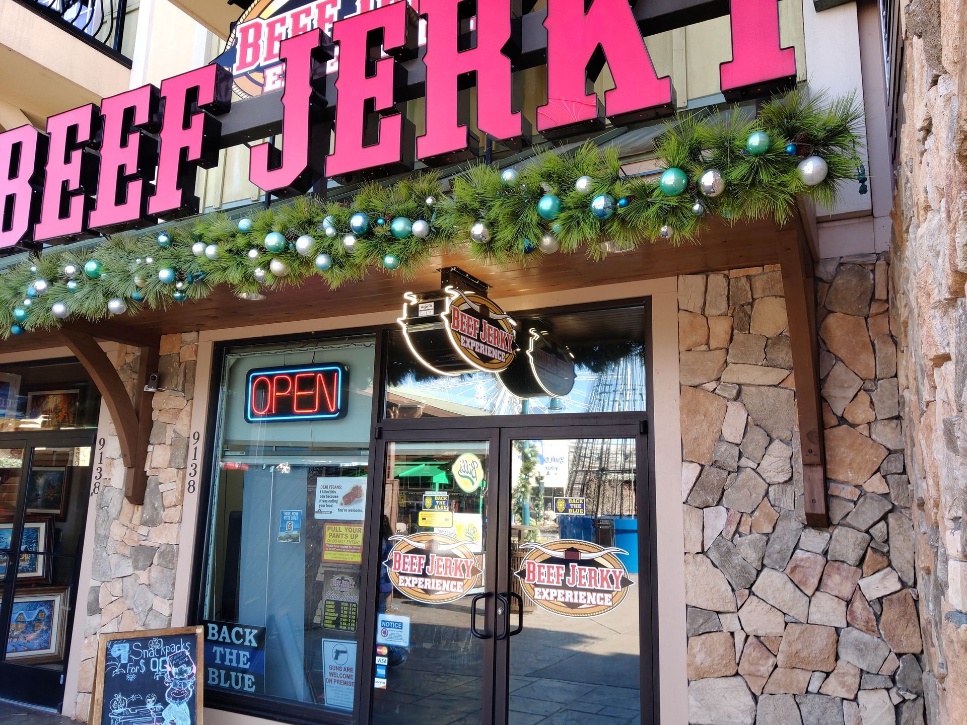 Storefront of Beef Jerky shop with pink sign, holiday garland, and neon OPEN sign