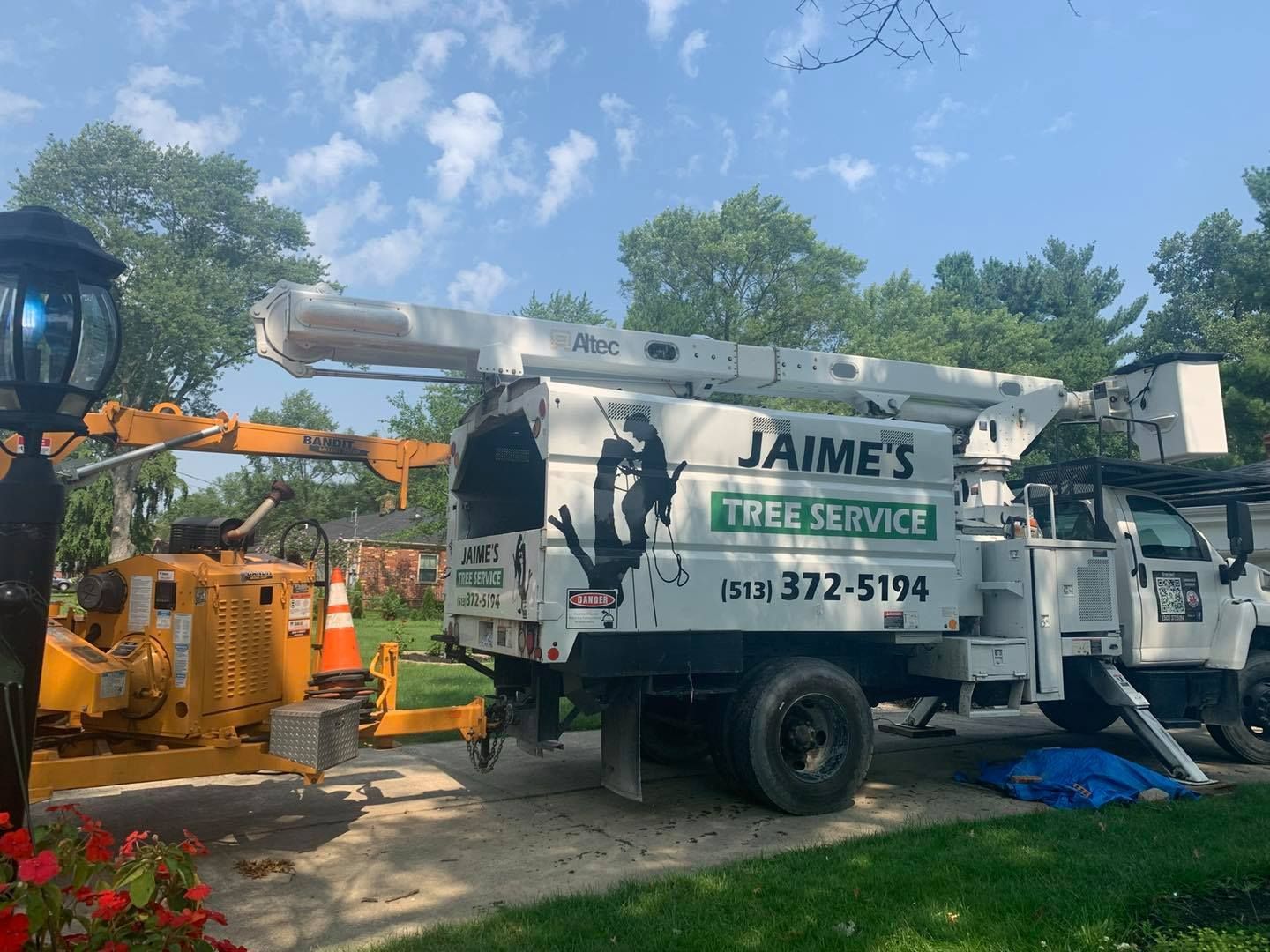 Jaime's Tree Service truck with a lift arm extended, next to a stump grinder. Sunny day, trees in the background.