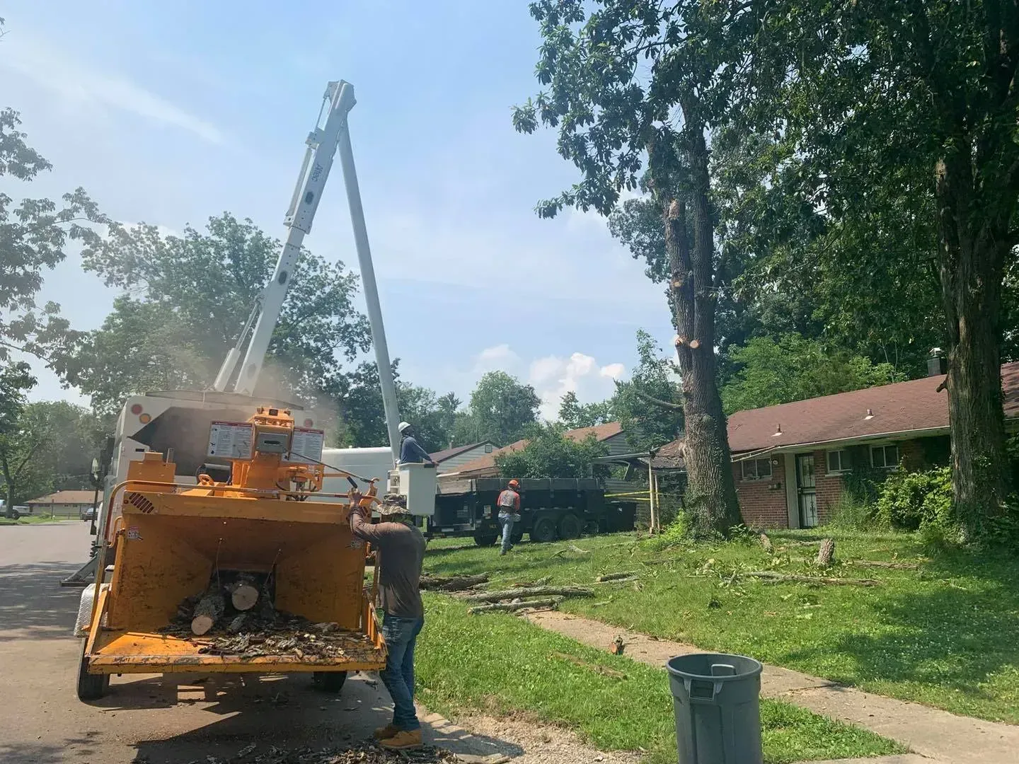 Tree service workers use a wood chipper and a boom lift to remove branches from a tree in a residential area on a sunny day.