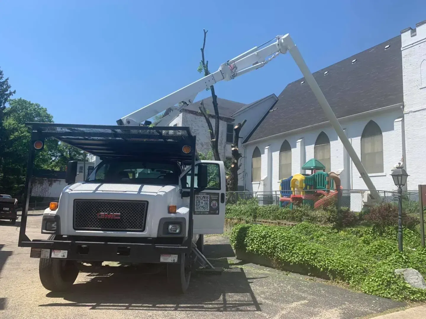 A tree service truck with its boom extended is trimming a tree near a white church building.  A playground is in the background.