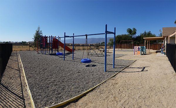 Playground with slides, swings, and climbing bars. Ground is covered in wood chips, fenced on two sides.