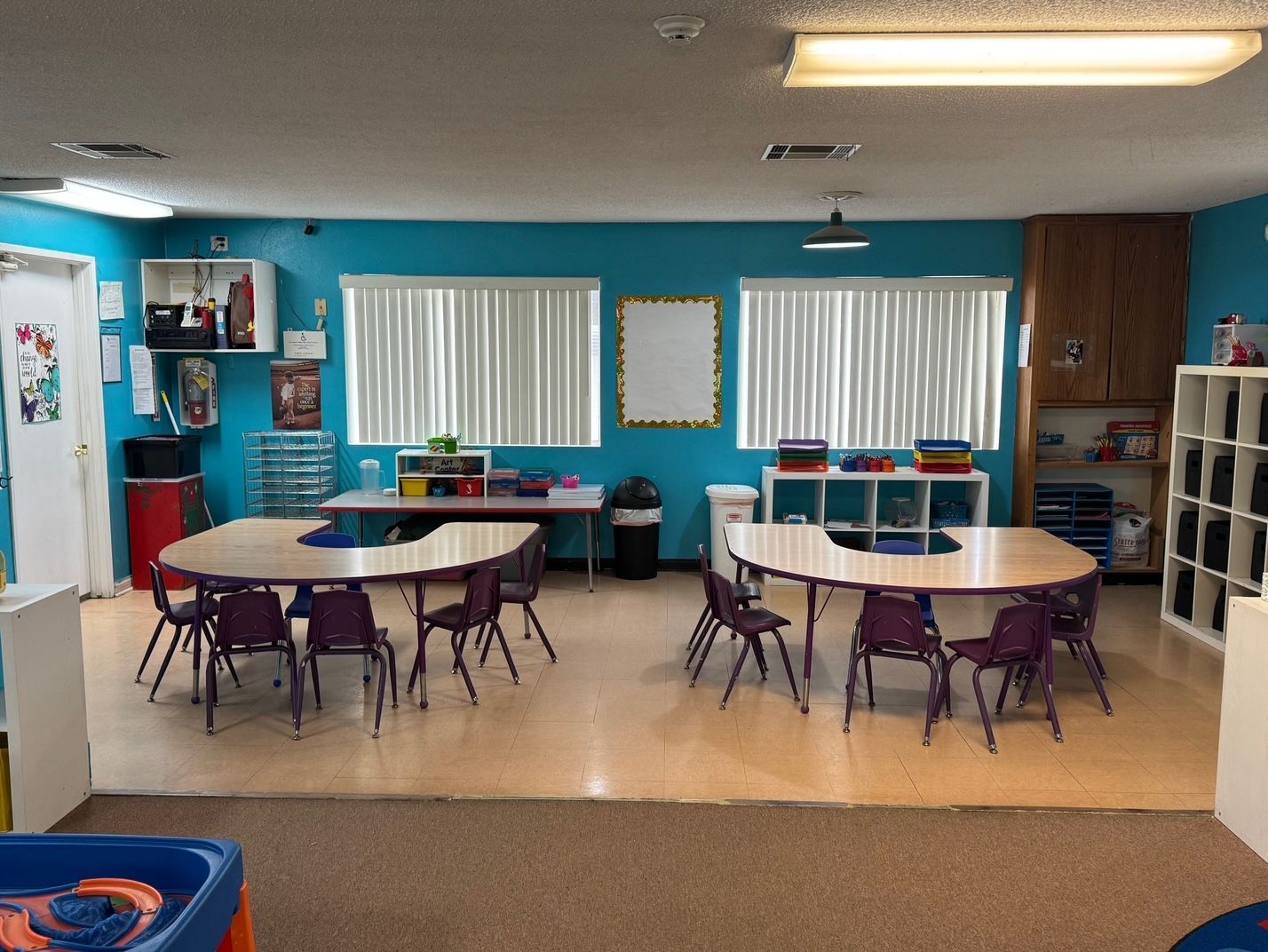 A classroom with oval tables, small purple chairs, windows with blinds, and blue walls.