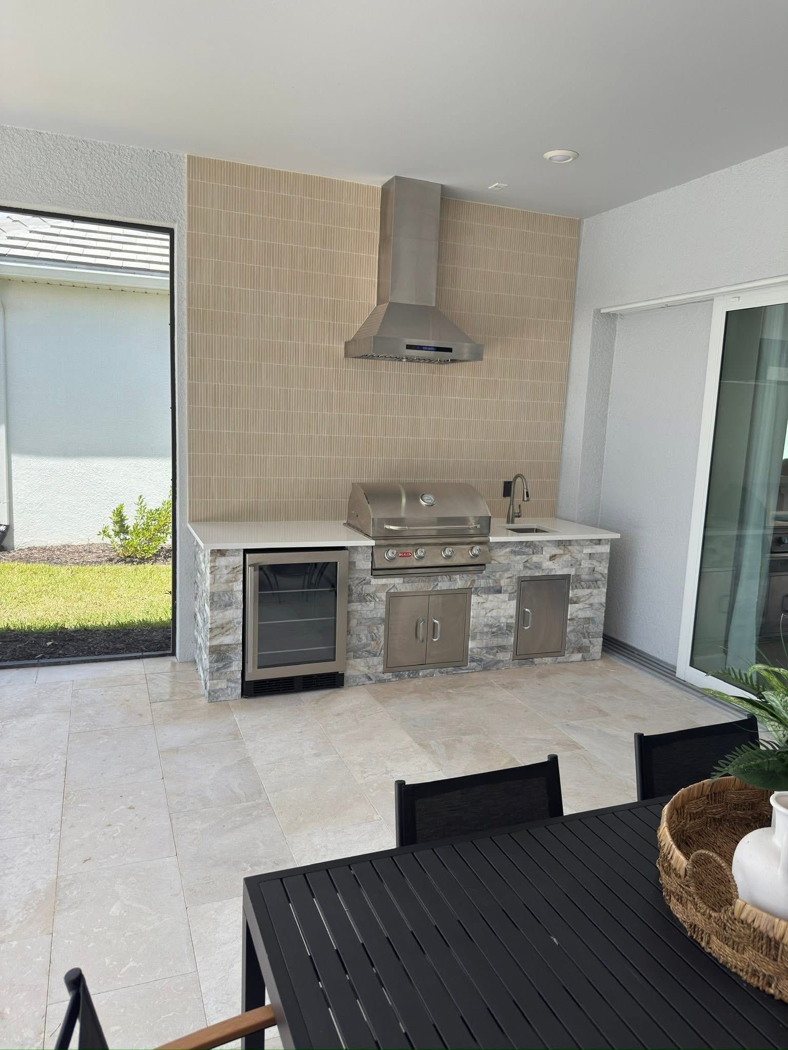 Outdoor kitchen with grill, vent hood, and beverage cooler, beige and gray tones.