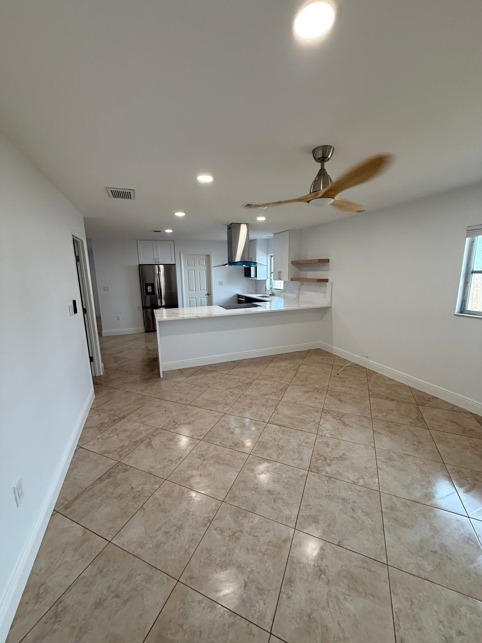 Empty, modern kitchen and living space with tile floors, white cabinetry, and a ceiling fan.