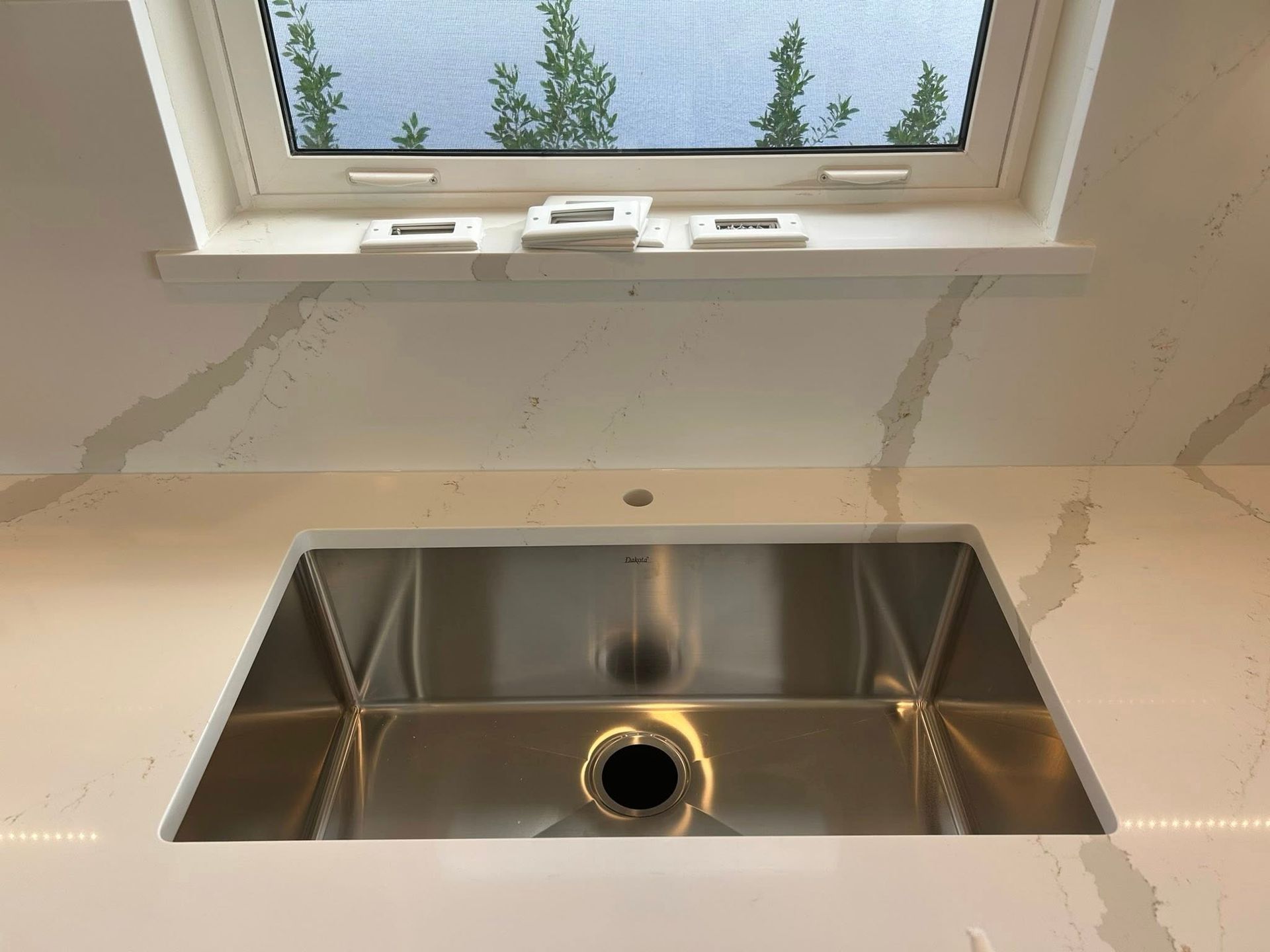 Stainless steel kitchen sink installed in a white countertop under a window.