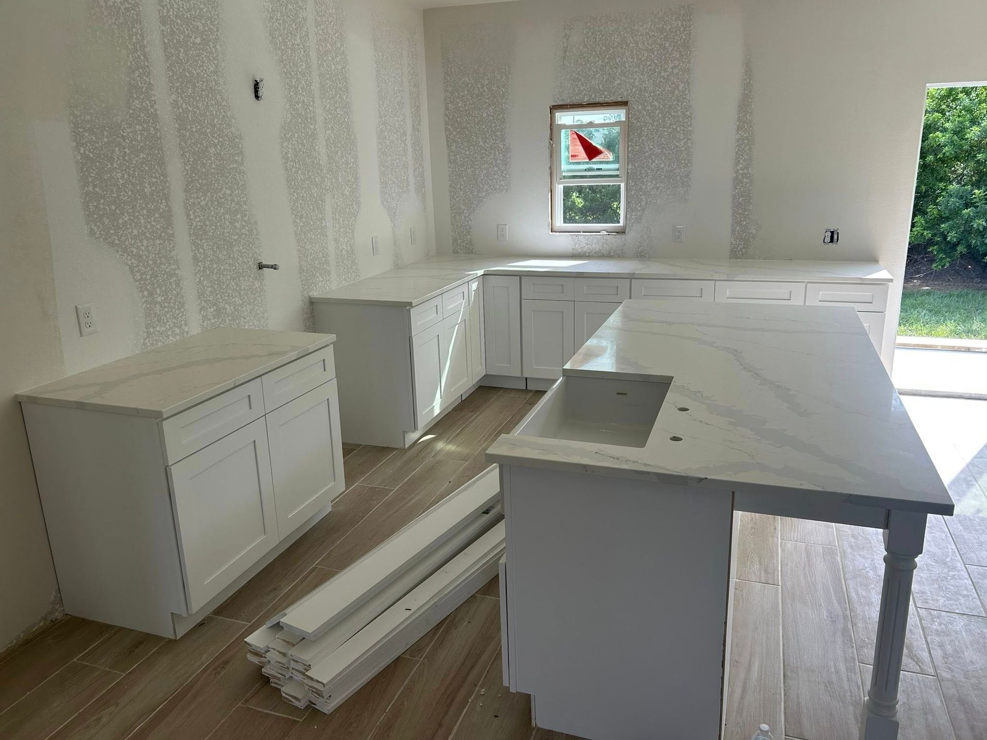 Kitchen under construction: white cabinets, countertops, and drywall. Flooring and trim are partially installed.