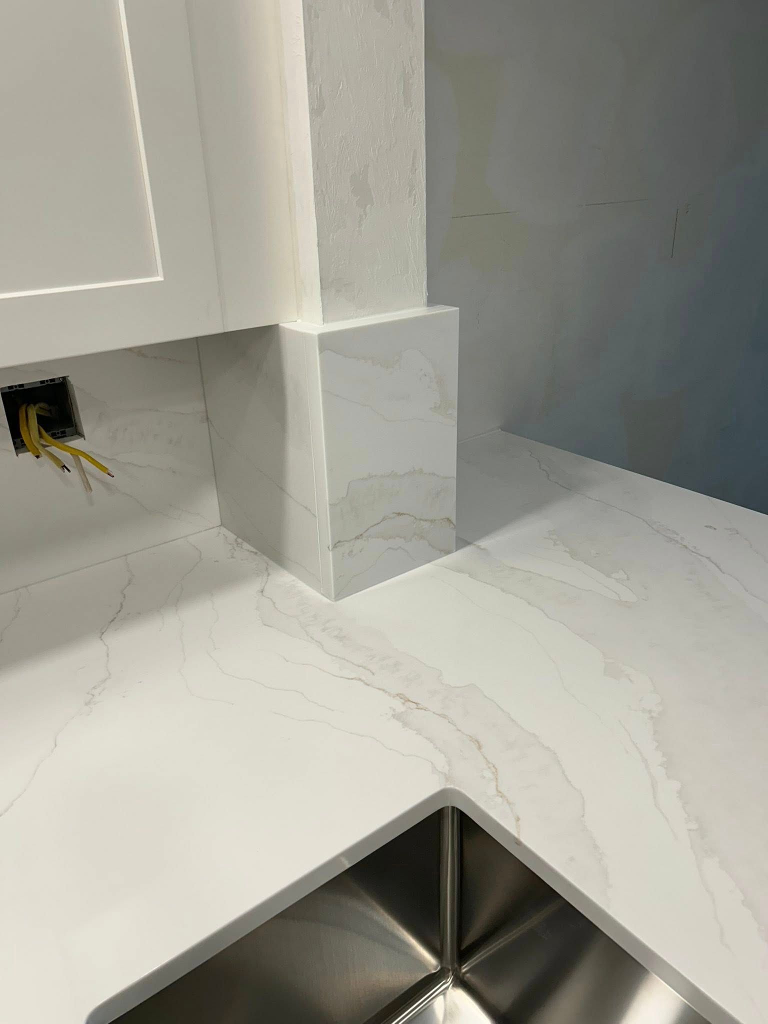 White quartz countertop and backsplash with a stainless steel sink in a kitchen.