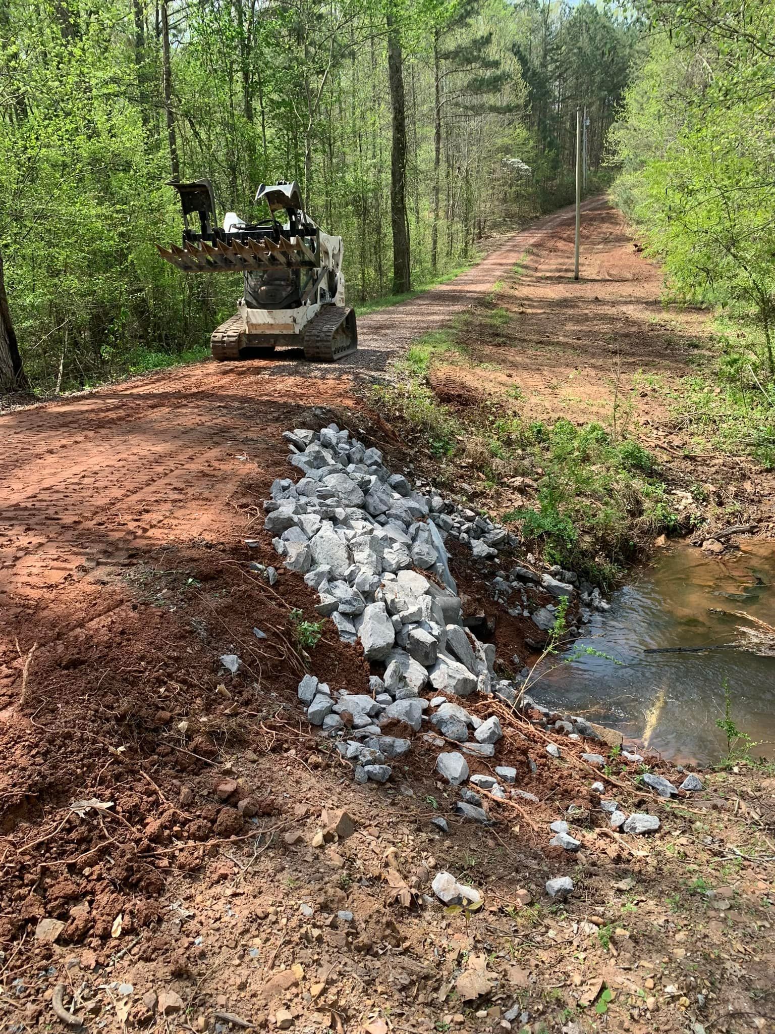 A bulldozer is driving down a dirt road next to a stream.