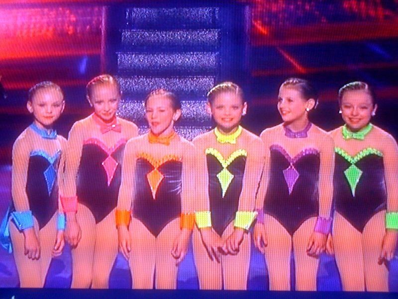Six gymnasts in colorful leotards posing on a stage with purple and red lighting