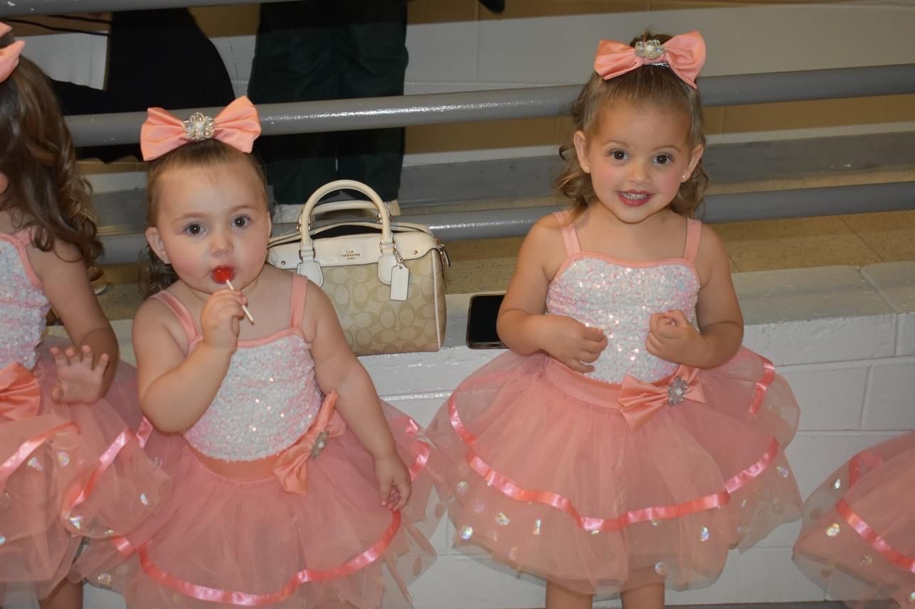 Two toddlers in pink ballet dresses and bows sitting on a bench, one sucking a lollipop.