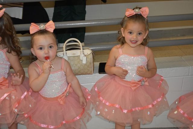 Two children in pink ballet dresses sitting on a bench, one sucking a lollipop and one smiling