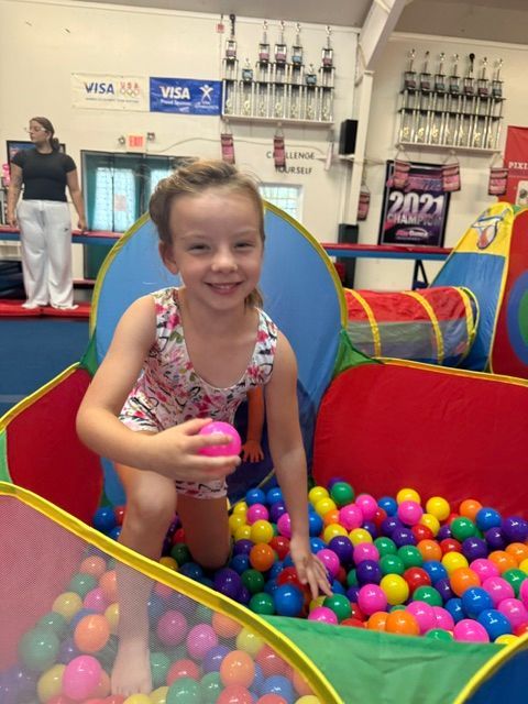 Child smiling in a colorful ball pit, holding a pink ball, with gym mats and equipment in the background