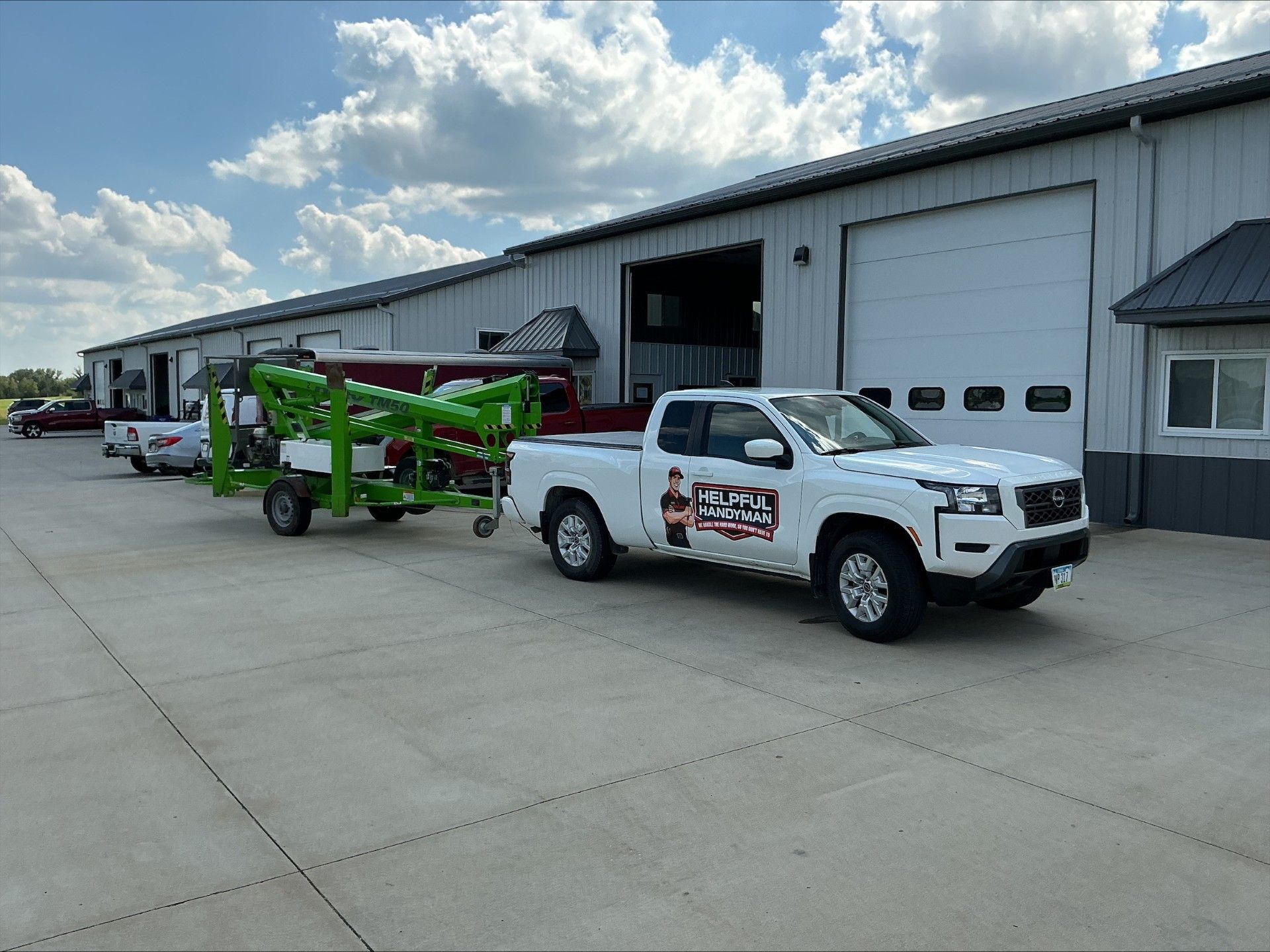 White truck towing a green trailer with equipment, parked in front of a gray industrial building.