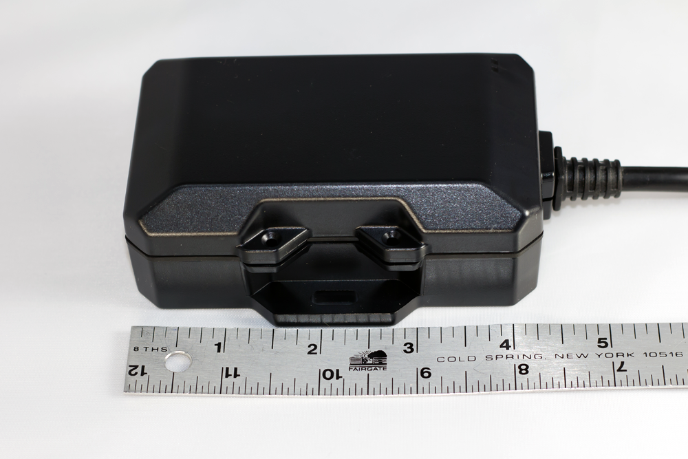 Black GPS tracker next to a ruler, showing size, on a white surface.