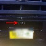Close-up of a black car bumper with an arrow pointing to a circular feature, and a license plate.