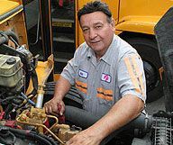 Mechanic working on a yellow bus engine, wearing a gray shirt with orange stripes.