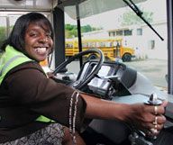 Smiling black woman driving a school bus, wearing a safety vest.