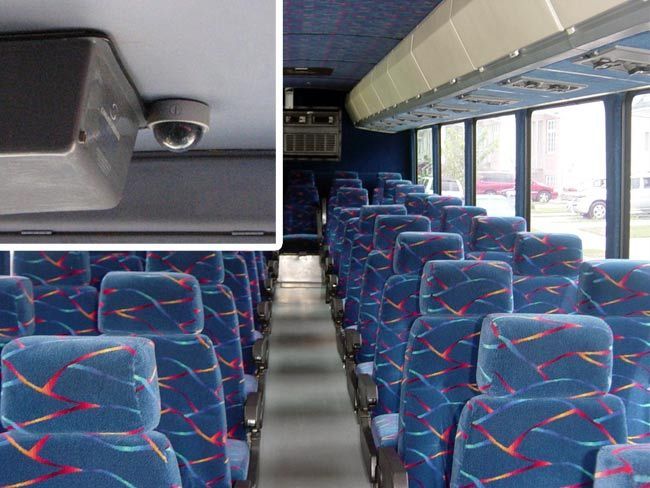 Interior of a bus with blue patterned seats and ceiling-mounted security camera; windows show outdoor setting.