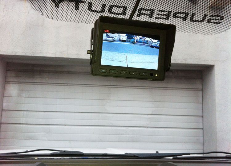 Backup camera display on a white wall above a garage door, showing a parking lot.