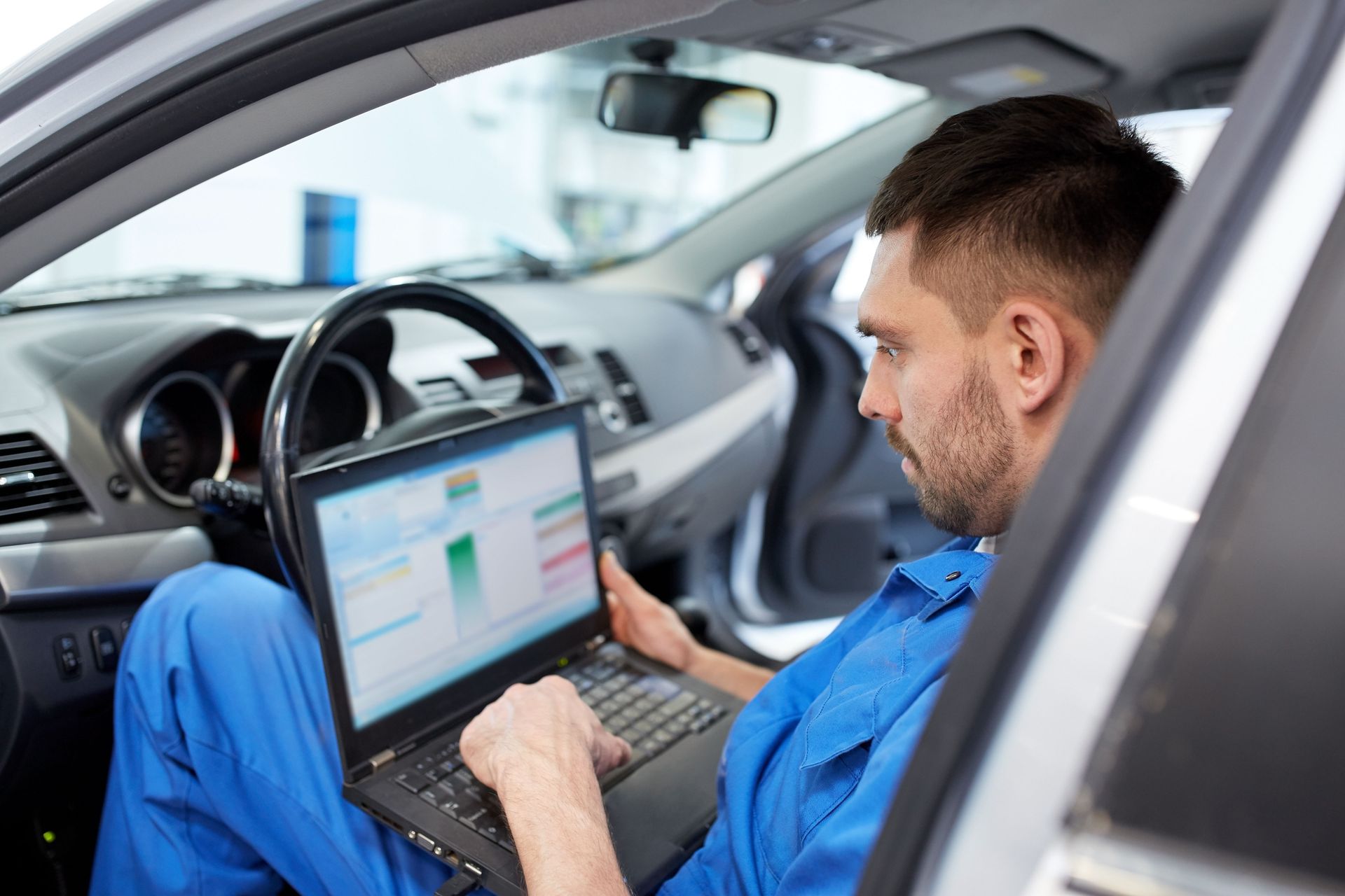 A mechanic in blue overalls using a laptop inside a car, likely diagnosing issues.