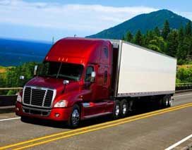 Red semi-truck driving on a highway, with a trailer. Ocean and mountains in the background.