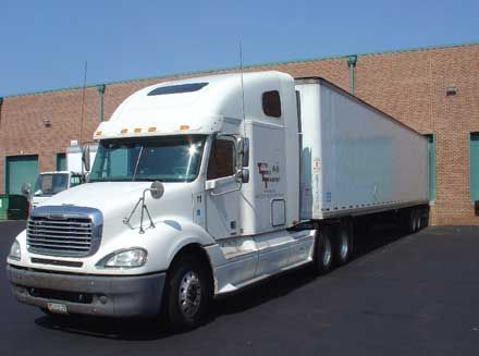 White semi-truck with trailer parked near a brick building on a sunny day.