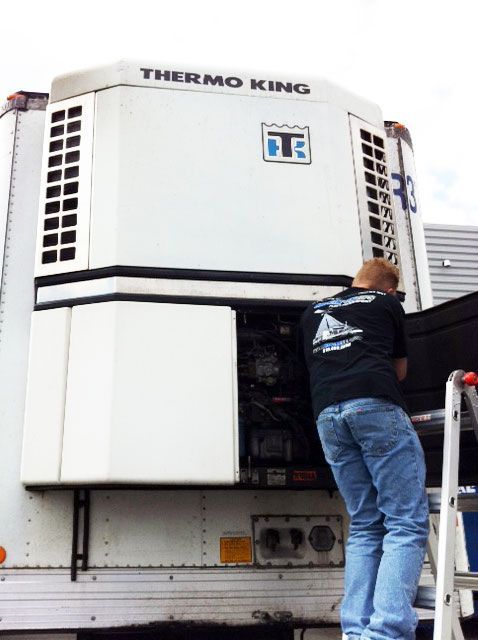 Man in jeans repairs a Thermo King refrigeration unit on a truck trailer, outdoors.
