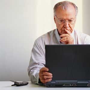 Elderly man in glasses, looking intently at a laptop screen, indoors, with a phone on the desk.
