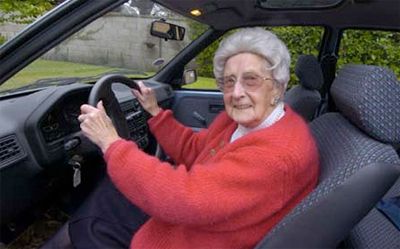 Elderly woman with white hair, red sweater, smiling, driving a car.