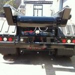 Rear view of a black garbage truck, showing the lift mechanism and tail lights, parked outdoors.