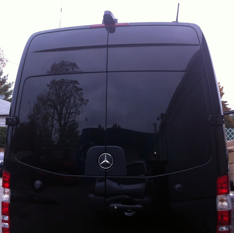 Black Mercedes Sprinter van rear view, tinted window reflecting trees and buildings.