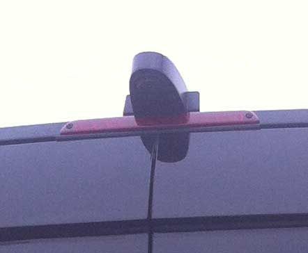 Backup camera above a vehicle's brake light; black camera, red light, dark gray vehicle.