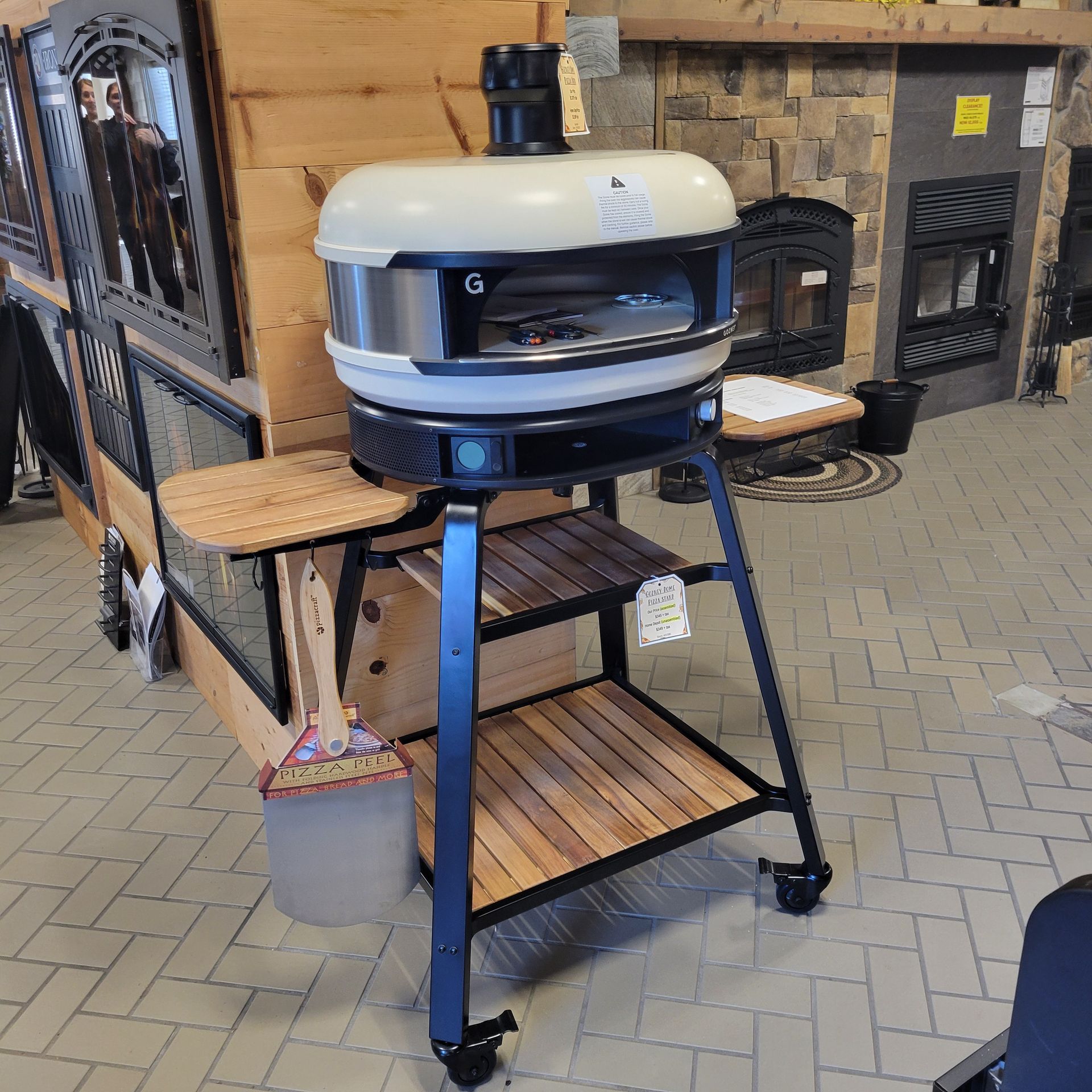 A pizza oven on a wheeled black stand. The oven is cream-colored, with a black chimney and viewing window.