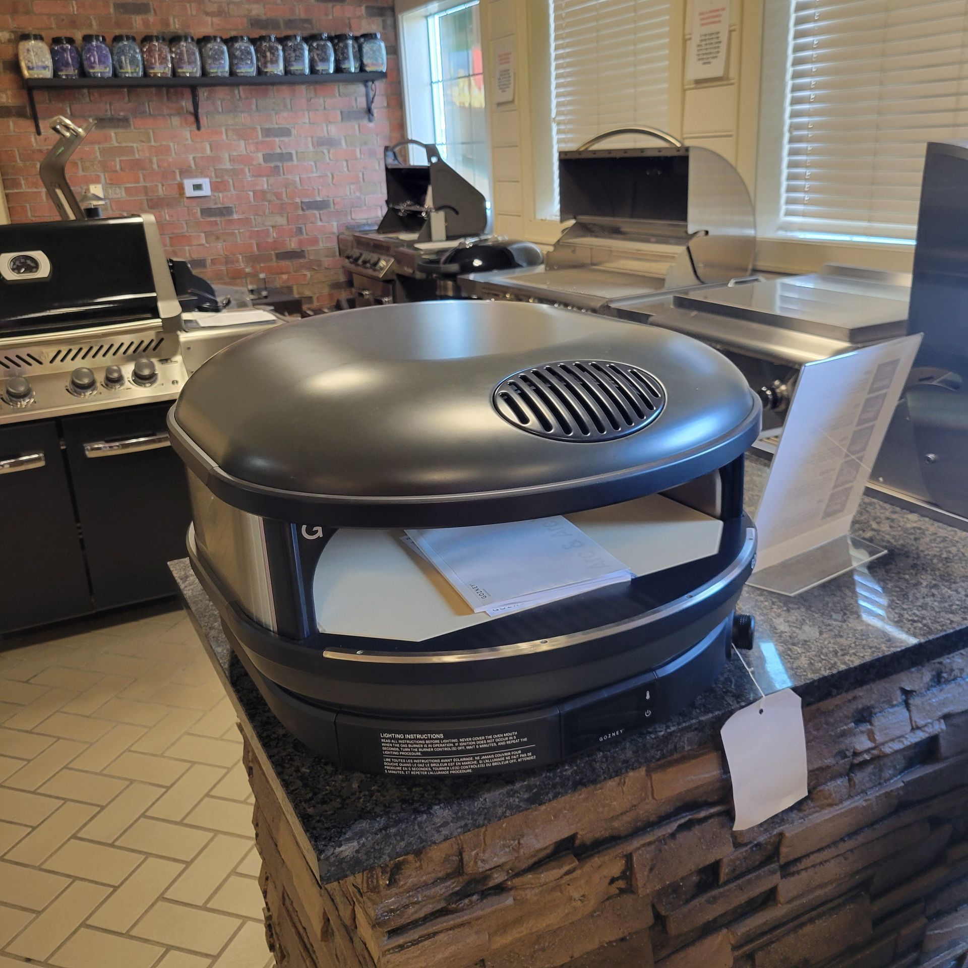 Black outdoor pizza oven on a counter in a showroom, with other appliances and shelves in the background.